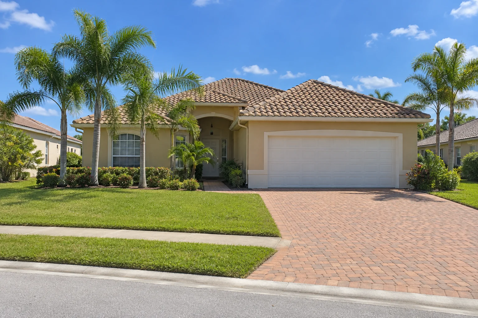 Single-story tan stucco home with a tile roof, paver driveway, and queen palms.